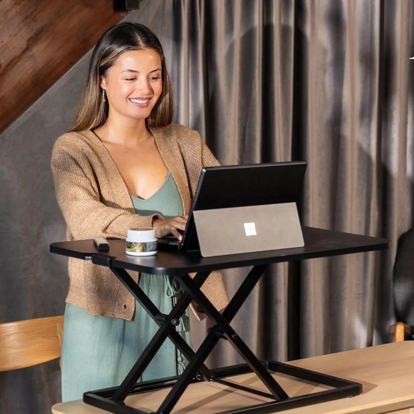 A woman in a casual outfit working on a laptop at a standing desk converter in a stylish indoor setting.