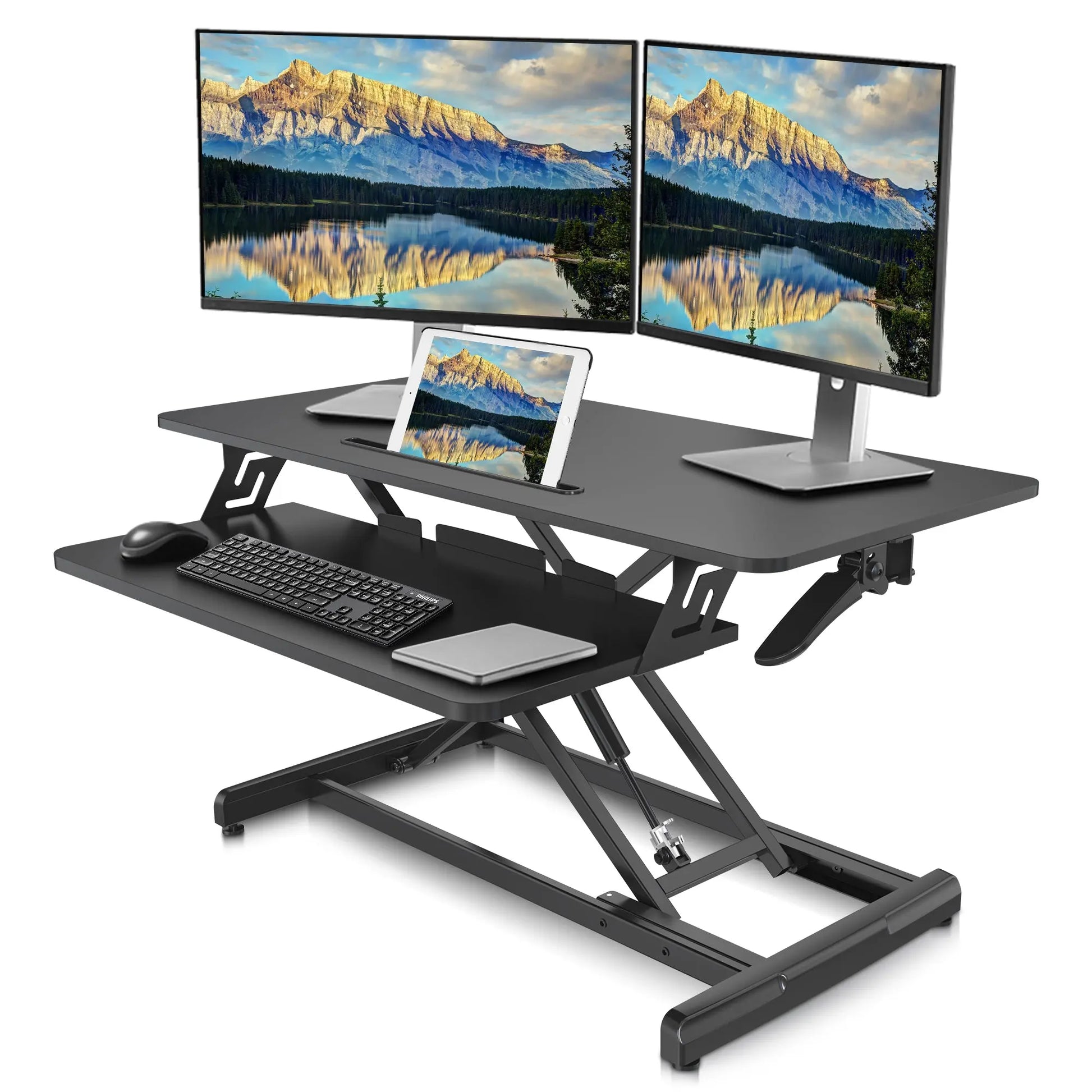 Dual monitor setup on a black sit-to-stand desk converter showcasing an expansive scenic wallpaper on screens, with keyboard, mouse, and tablet on the lower tier.