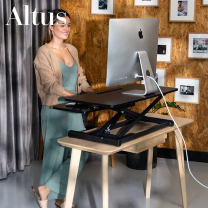 Woman standing at a modern workspace using a black adjustable desk riser with a desktop computer; stylish office with wood paneling and framed photos in the background.