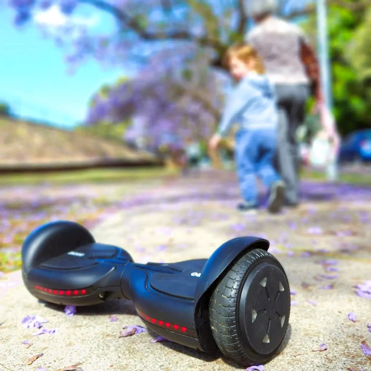 This image shows a black hoverboard placed prominently on a pathway covered with fallen purple flowers, likely from jacaranda trees. In the background, slightly blurred, a child and an adult are seen walking away, giving a sense of family activity and outdoor enjoyment. The focus on the hoverboard in the foreground with the active, familial background suggests a lifestyle where technology meets leisure and outdoor fun