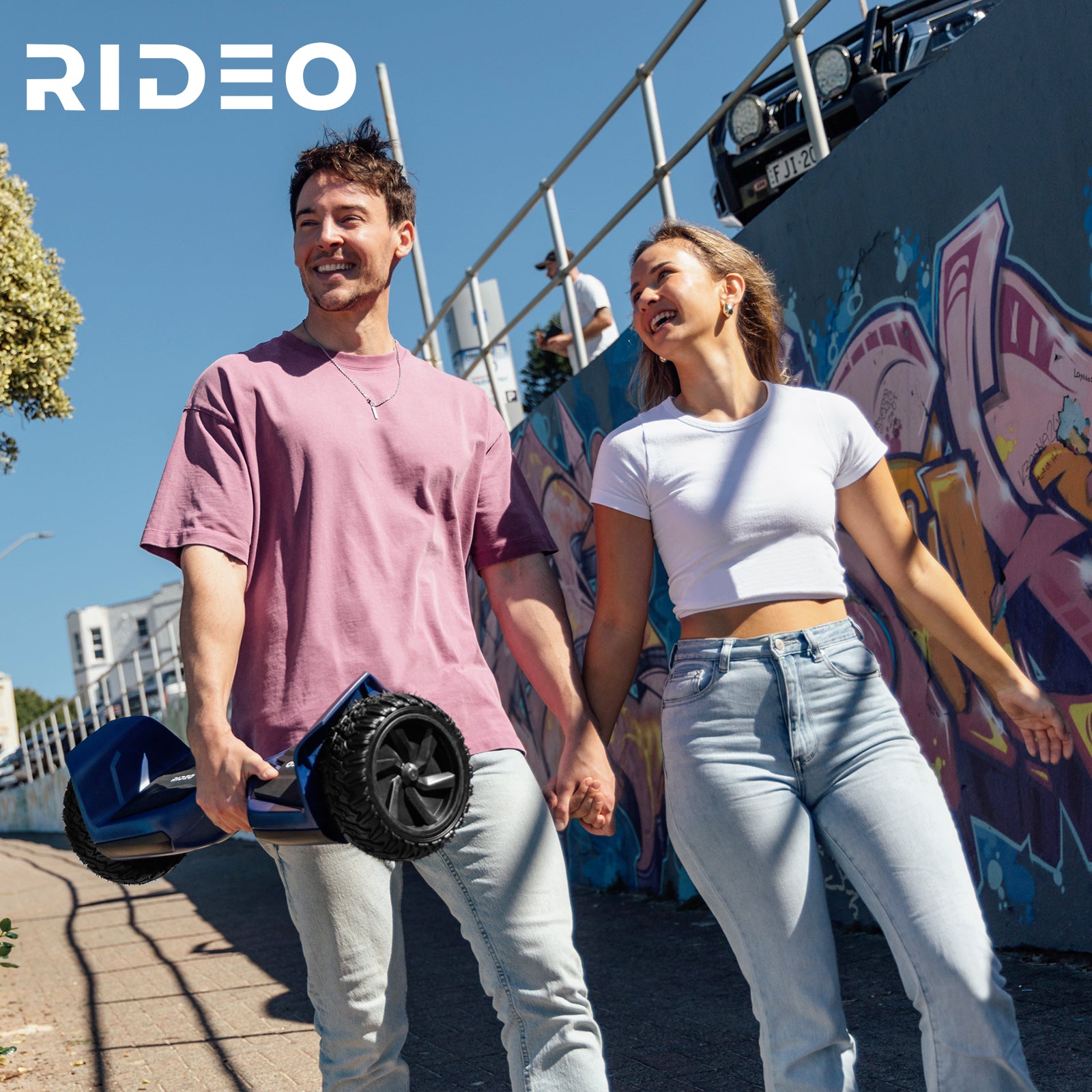 A young couple enjoying a sunny day outdoors, holding hands while one of them carries a blue Rideo hoverboard. They are walking along a graffiti-covered wall, symbolizing a vibrant and dynamic lifestyle.