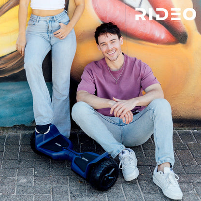 A young man sitting next to a blue Rideo hoverboard in an urban environment, enjoying a relaxed moment. The image conveys a modern and adventurous lifestyle.