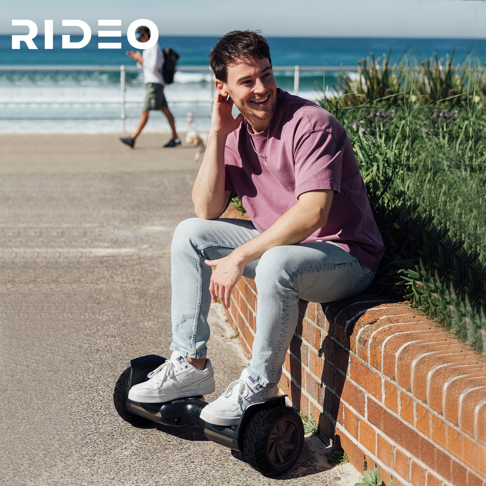 A young man sitting comfortably on a Rideo hoverboard by a scenic beachside, enjoying a sunny day. The image captures a carefree and adventurous lifestyle, highlighting the hoverboard's portability and versatility in various environments. Perfect for promoting the fun and freedom of Rideo hoverboards in outdoor settings.