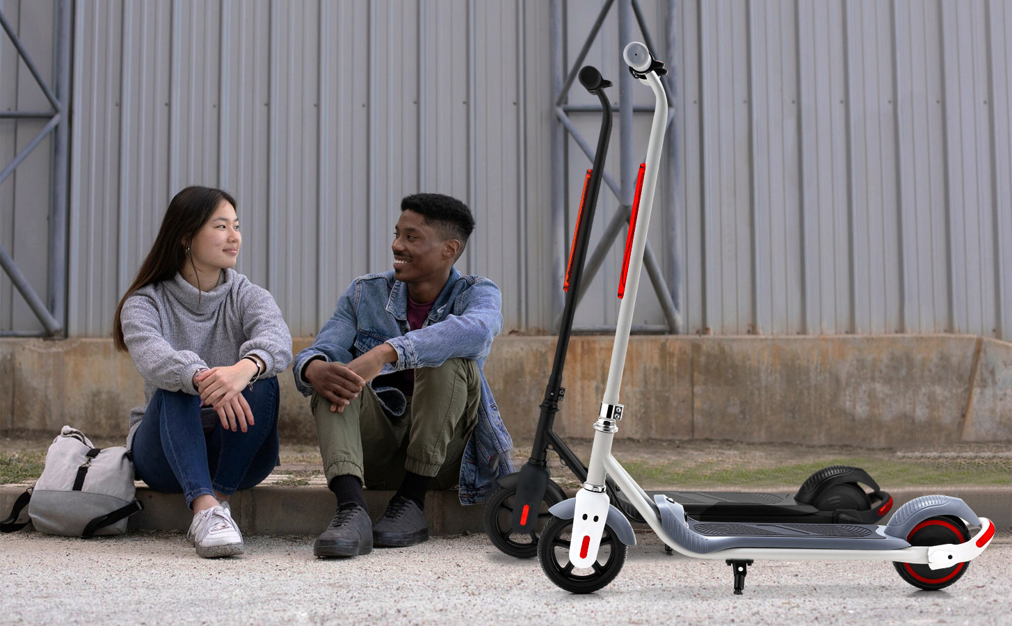 Lifestyle image of a young woman and man seated on the ground next to the white electric scooter, chatting in an urban setting.