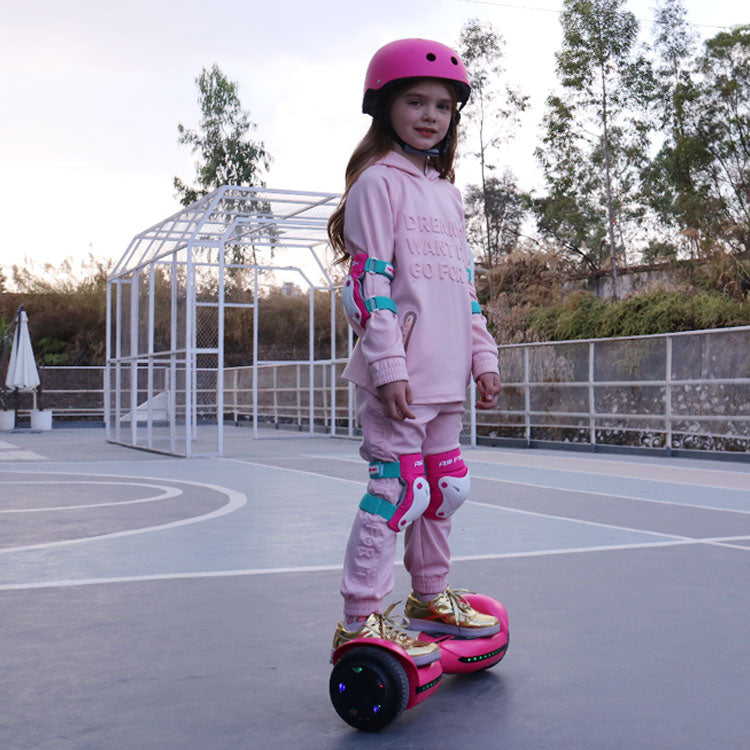 A young girl in a pink helmet and matching outfit stands on a vibrant pink and gold hoverboard equipped with blue LED lights. She's in a basketball court setting, exuding confidence and style, which highlights the hoverboard's appeal to younger users.