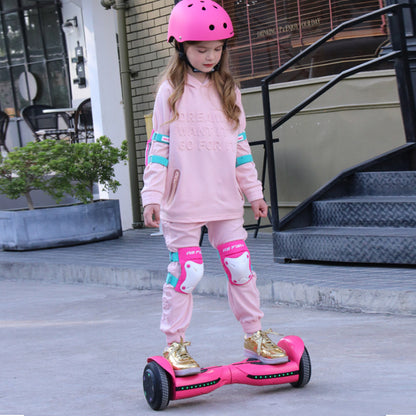 A young girl in a pink helmet and matching outfit stands on a vibrant pink and gold hoverboard equipped with blue LED lights. She's in a basketball court setting, exuding confidence and style, which highlights the hoverboard's appeal to younger users.